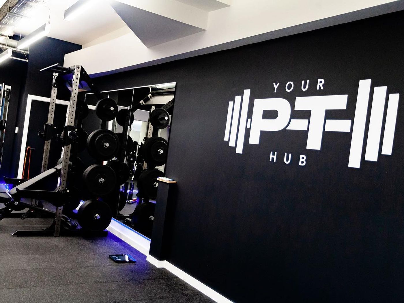 Power rack and free weights beneath Your PT Hub's illuminated wall logo, in the studio's mirrored training area