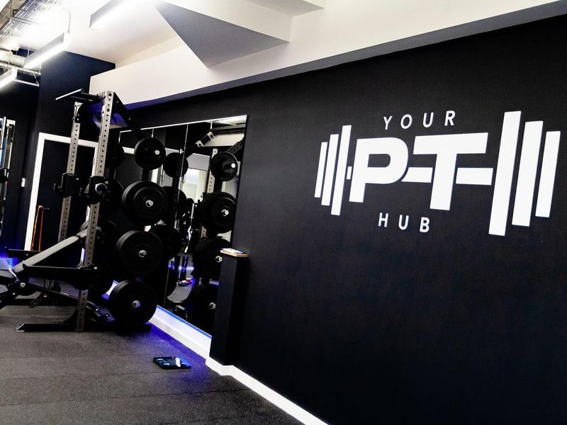 Power rack station beneath the studio's illuminated wall logo, Your PT Hub